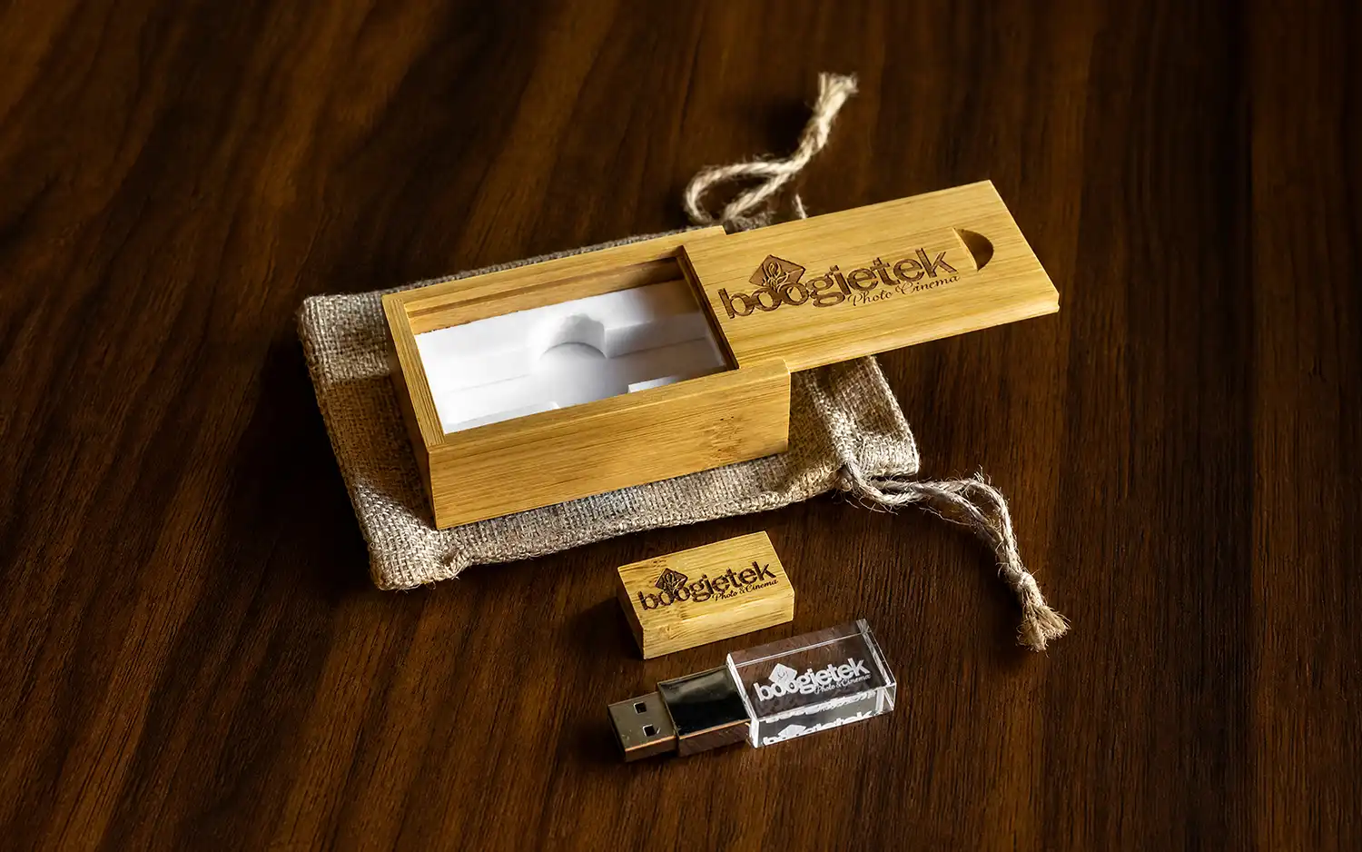 A boogietek USB drive box open on a burlap pouch on a dark wood table, beside a small engraved wood USB stick and a crystal USB drive, by Boogietek Photo+Cinema
