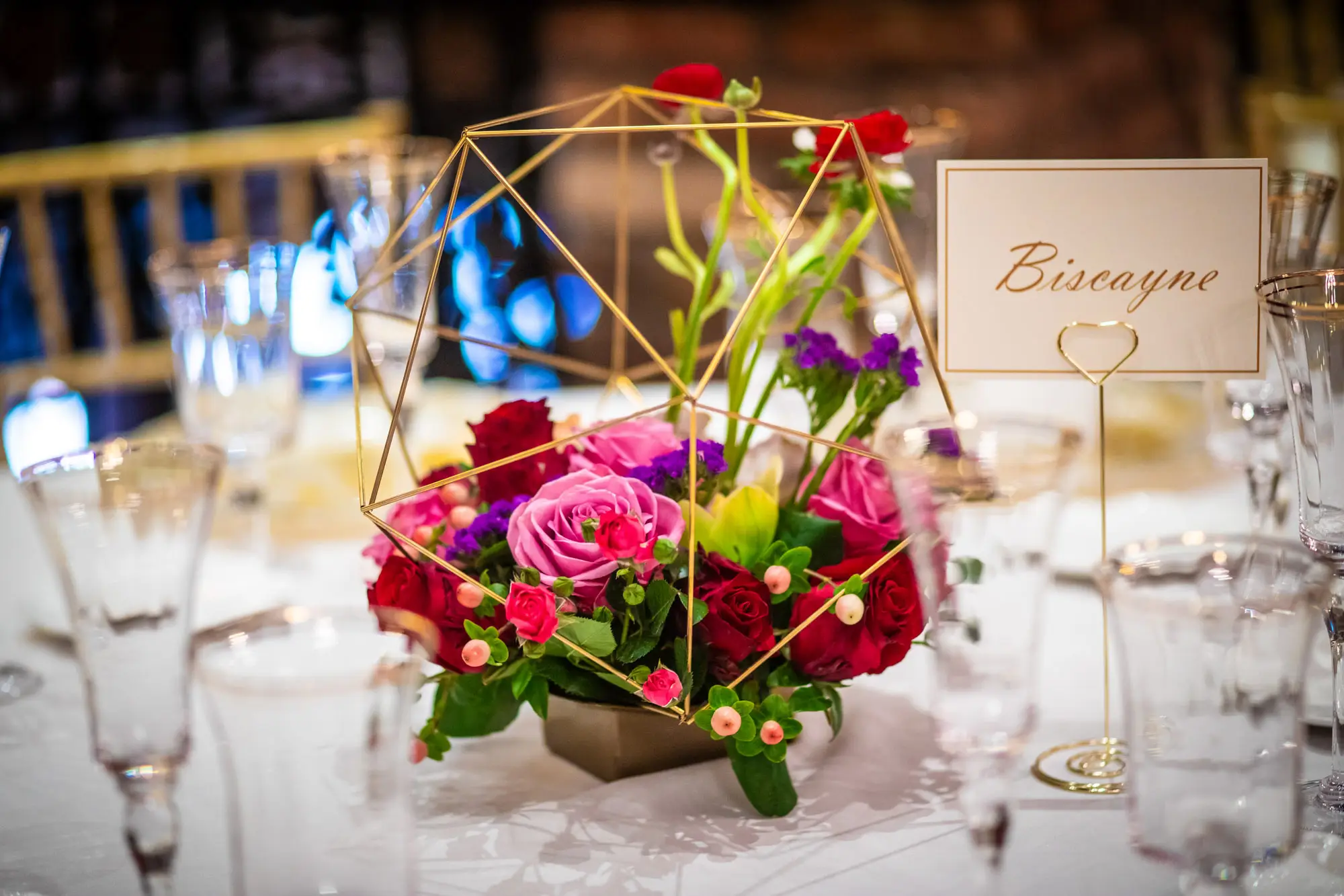 Reception centerpiece of pink and red roses in a gold geometric frame beside a Biscayne table card at The Cruz Building in Miami by Boogietek Photo+Cinema