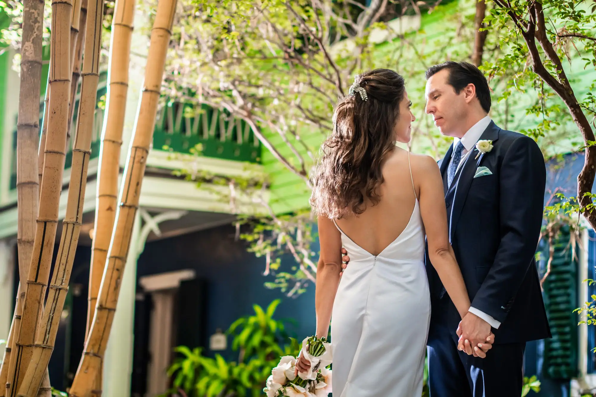 Bride in a backless white gown holding peonies and her groom in a navy suit sharing a quiet look beneath tall bamboo in a garden courtyard by Boogietek Photo+Cinema