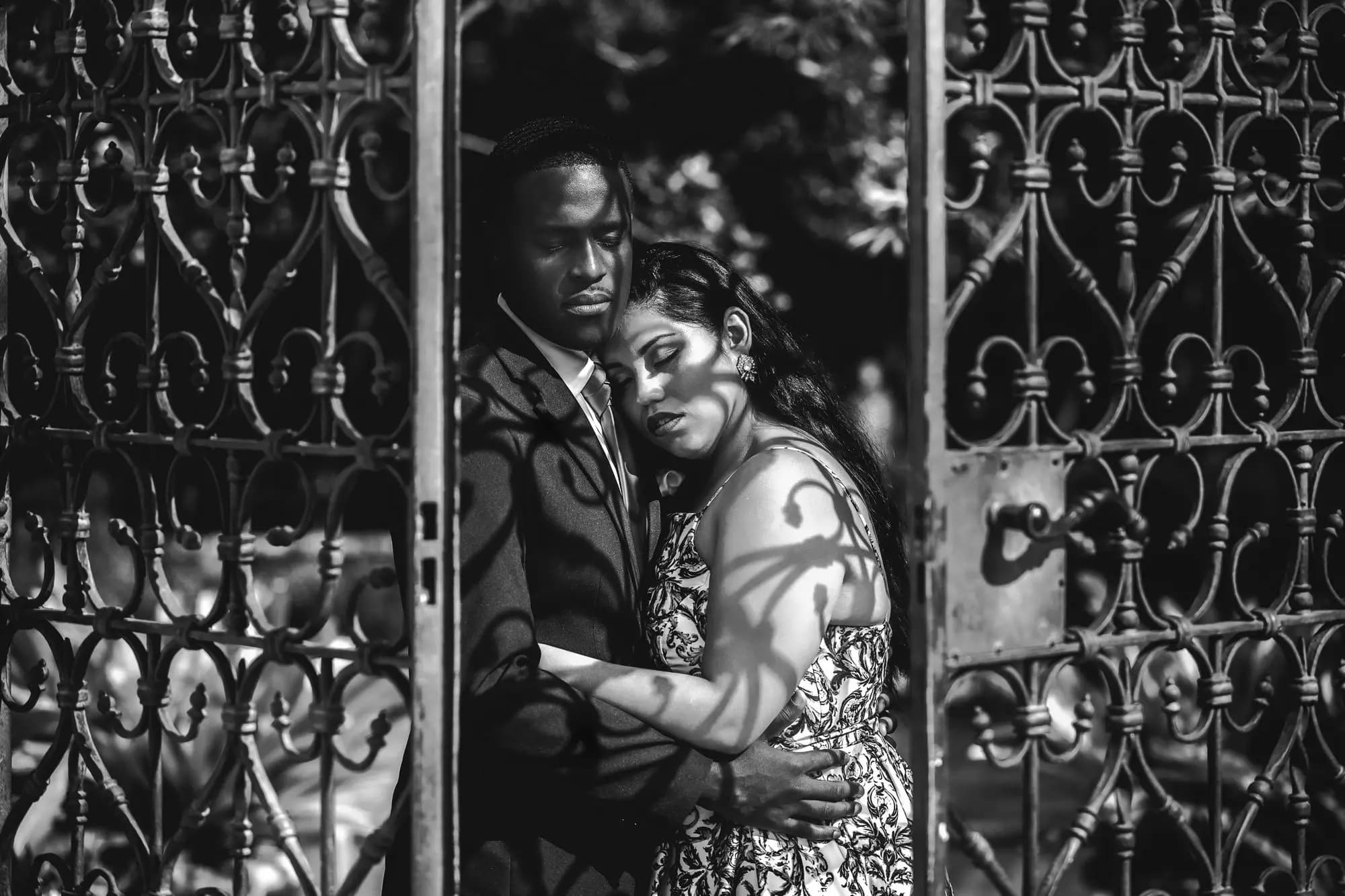 Bride and groom framed inside an ornate wrought-iron gate, foreheads together with eyes closed as dappled leaf shadows fall across them, by Boogietek Photo+Cinema