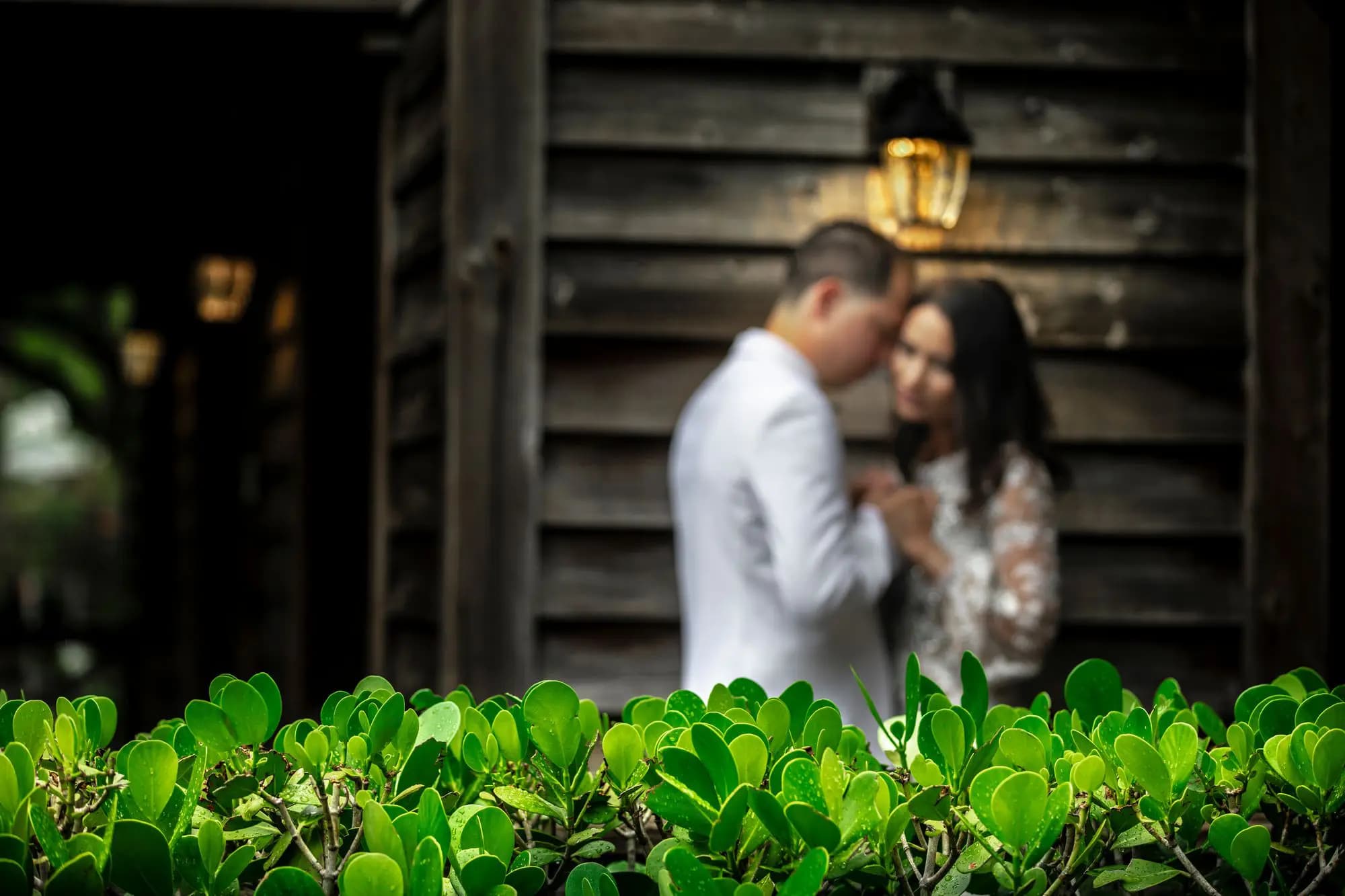 Groom in a white jacket and bride in a lace gown leaning together against weathered wood siding under a glowing lantern by Boogietek Photo+Cinema